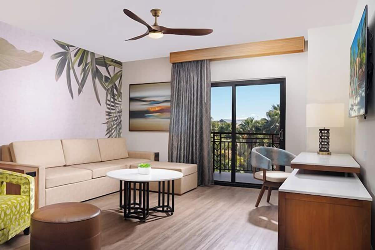 A modern hotel room with a beige sofa, a green patterned armchair, a round table, a ceiling fan, a balcony with a view, and a painting on the wall.