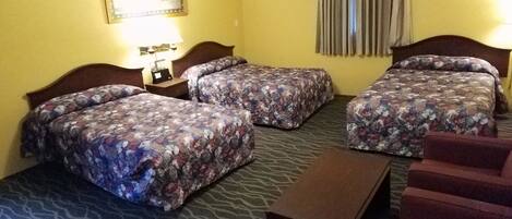 Habitación de hotel con tres camas, una mesita de noche con lámpara, una pequeña mesa y una silla.