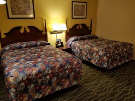 Habitación de hotel con dos camas, cada una con una colcha floral, una mesita de noche con lámpara y cuadros enmarcados en la pared.