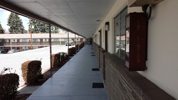 A covered corridor with a fire extinguisher on the wall, a parking lot with cars, and a building with a red roof in the background. - Budget Host Midway Inn (Bedford)