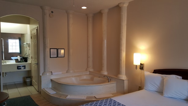 Un baño de hotel con una bañera grande, un lavamanos y un espejo. Hay toallas en el suelo y una lámpara en la pared.
