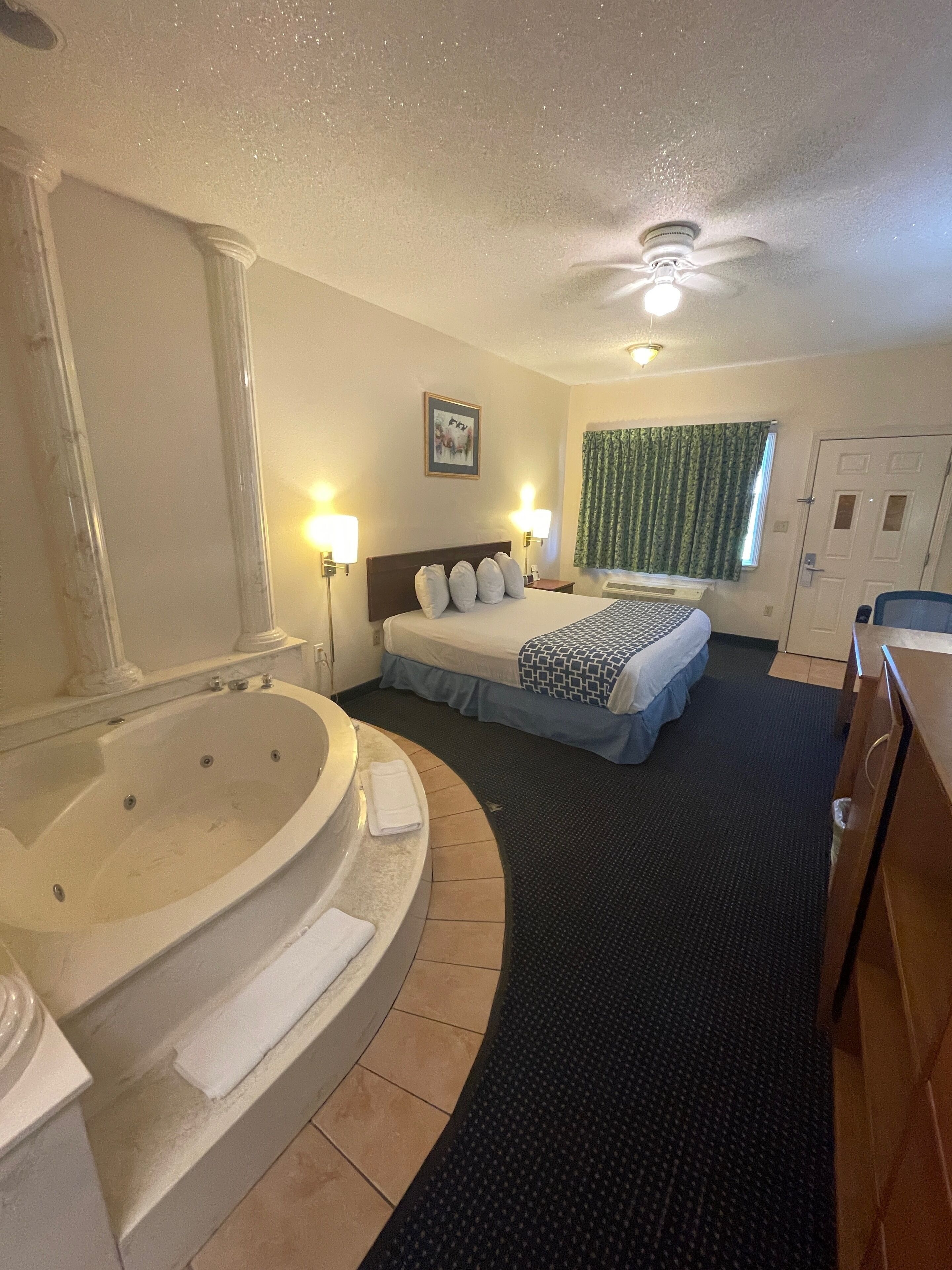 Habitación de hotel con una bañera grande, una cama doble con cubrecama estampado, dos lámparas de pared, un ventilador de techo y una ventana con cortinas.