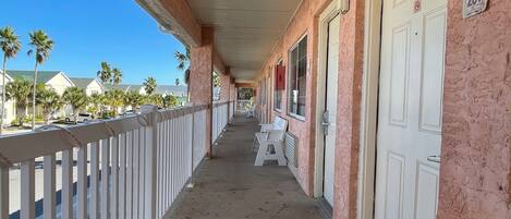 A hotel corridor with a covered walkway, white doors, and a row of white chairs.
