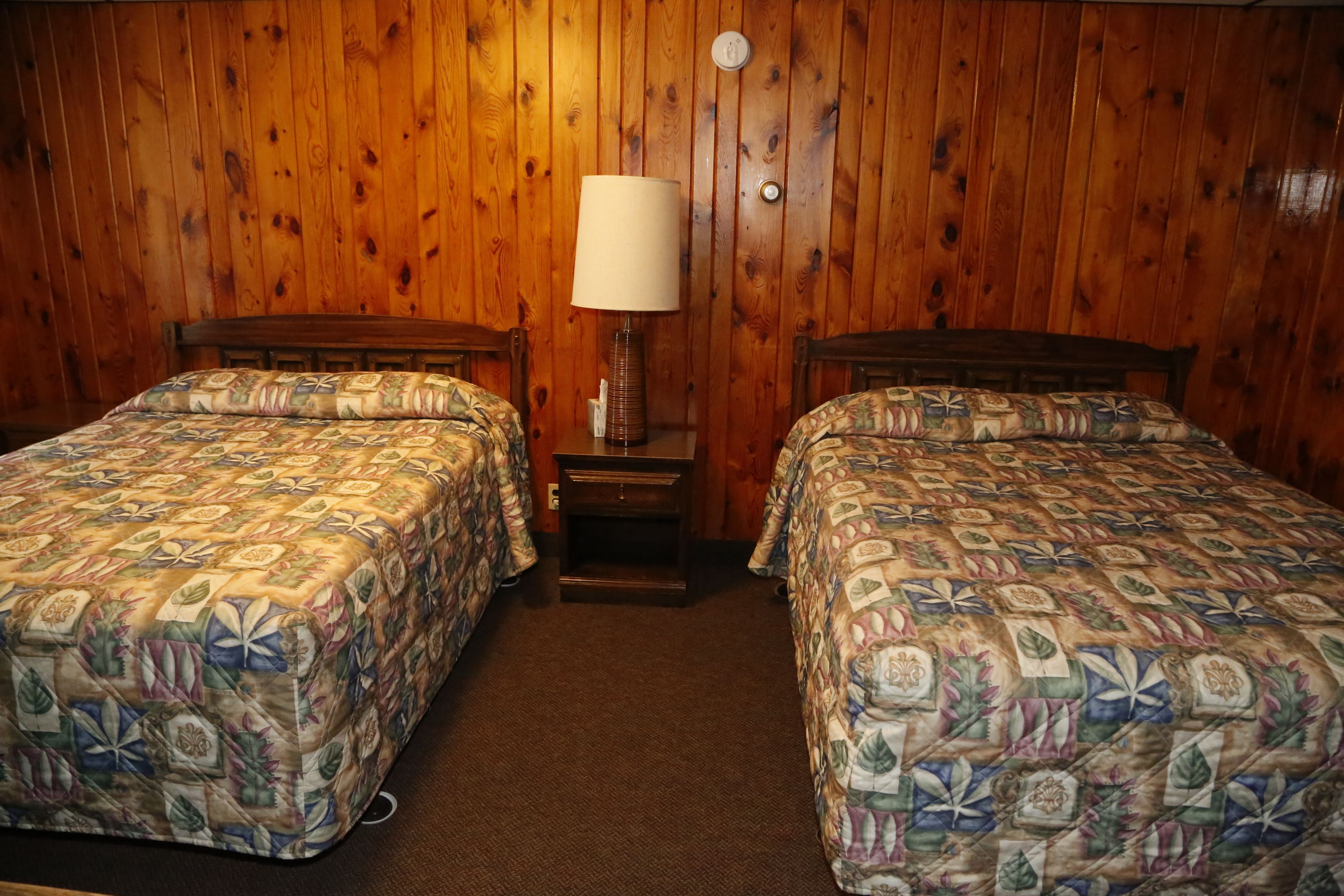 A room with two beds, wooden walls, a nightstand with a lamp, and a carpeted floor.