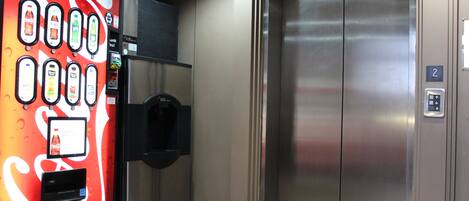 A vending machine with various drink options and a water dispenser, located next to a metallic elevator.