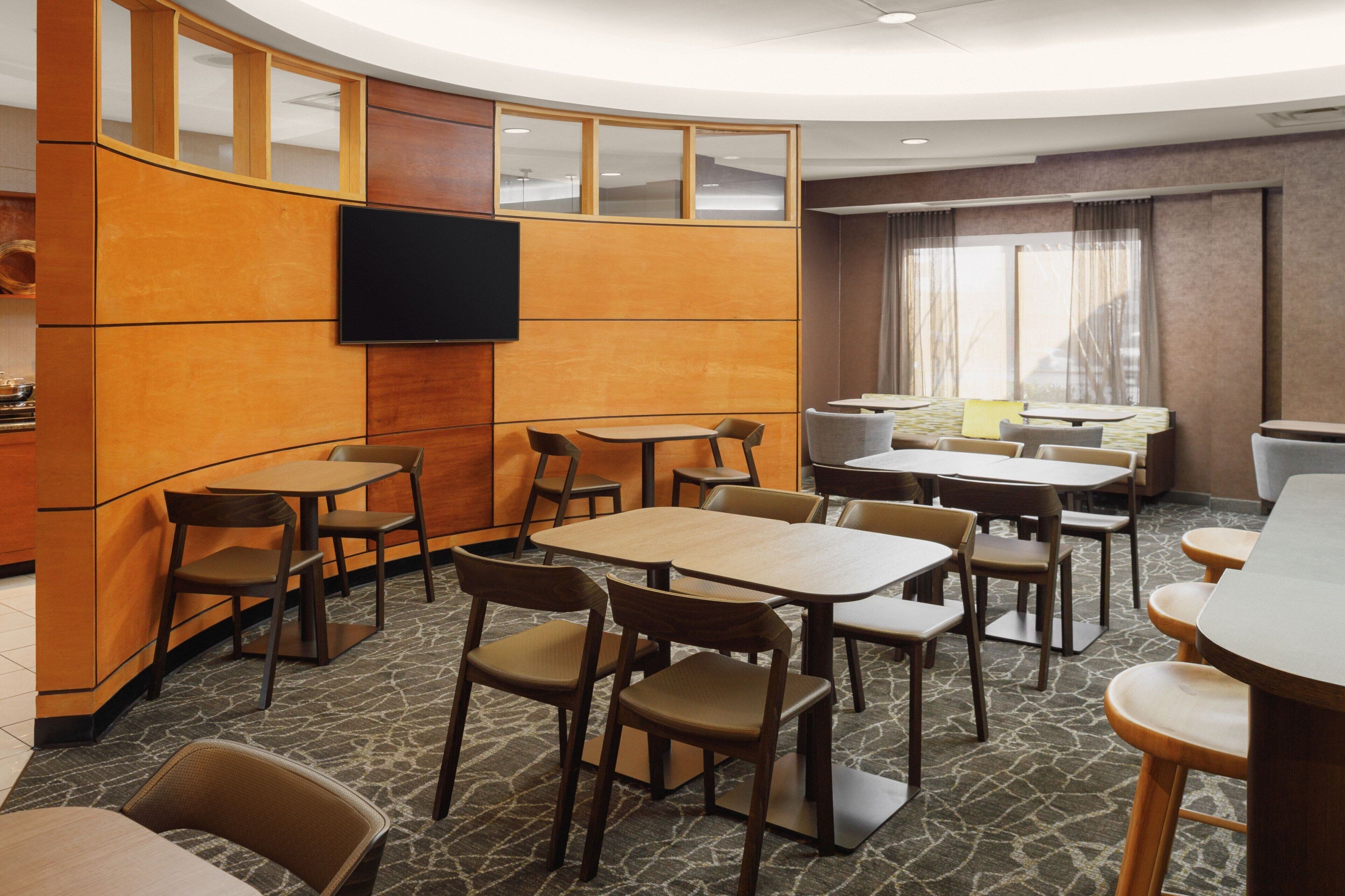 A hotel dining area with multiple tables and chairs, a flat-screen TV mounted on the wall, and a large window with curtains.
