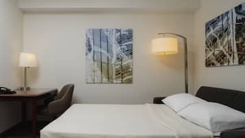 Habitación de hotel con cama, escritorio con lámpara, una silla y dos cuadros colgados en la pared.