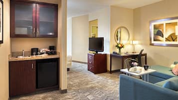 Quarto de hotel com cozinha compacta, escrivaninha com computador, televisão, sofá e espelho.