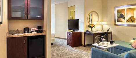 A hotel room with a kitchenette, a desk with a computer, a television, a sofa, and a mirror.