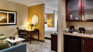 A hotel room with a bed, a desk with a lamp, a chair, a small table with a vase, and a kitchenette with a coffee maker and sink.