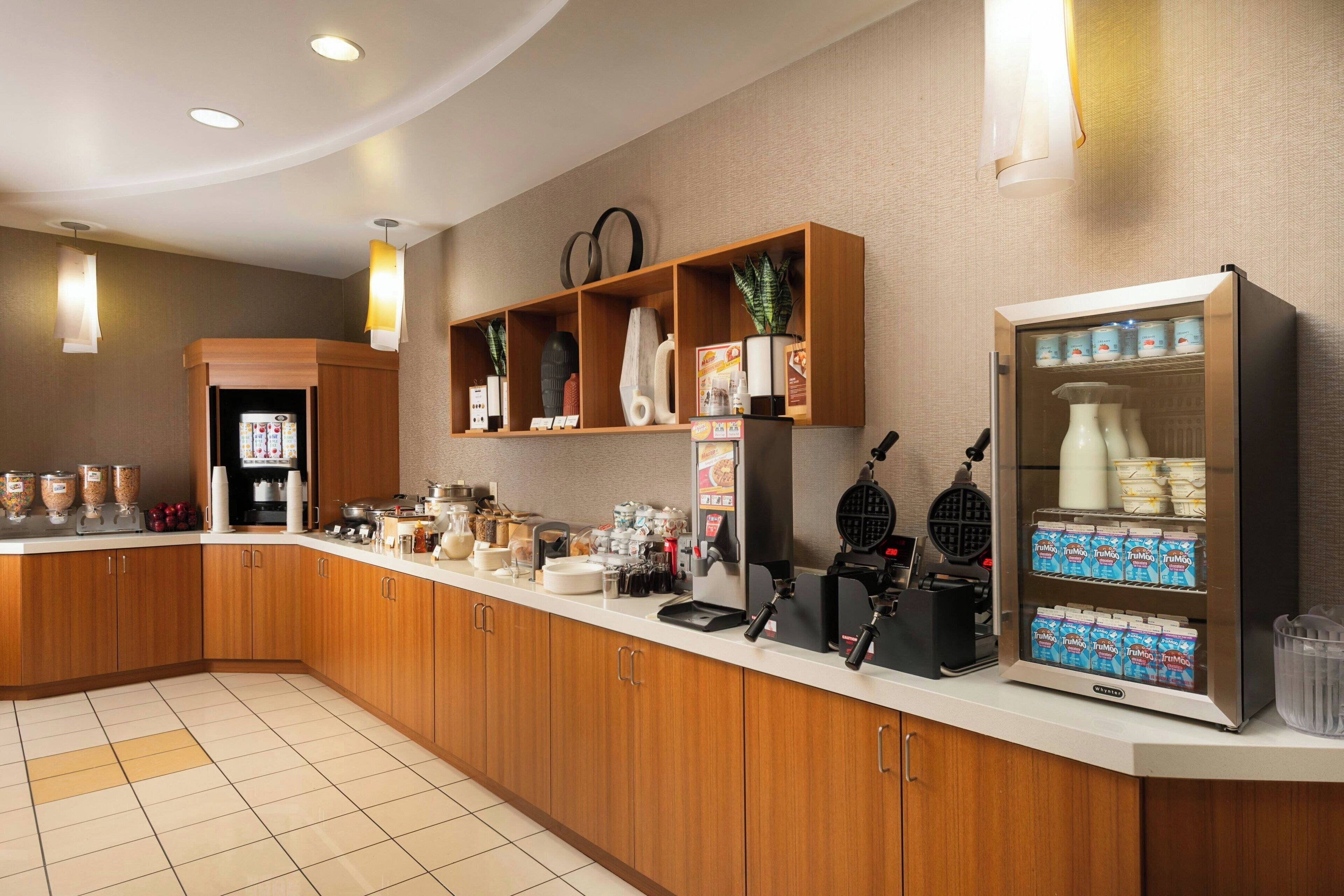 A hotel kitchen with wooden cabinets, a coffee machine, a waffle maker, and a refrigerator stocked with beverages.