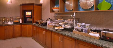 A hotel kitchen with wooden cabinets, a self-serve breakfast station, and decorative wall art.