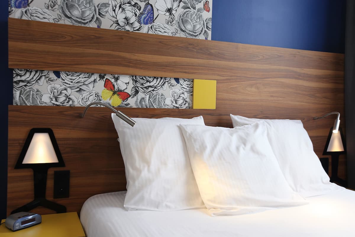 A neatly made bed with white pillows and a wooden headboard with floral wallpaper. There are two bedside lamps and a small table with a clock.