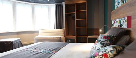 A modern hotel room with a large bed, a wooden wardrobe, a desk, and a window with curtains.