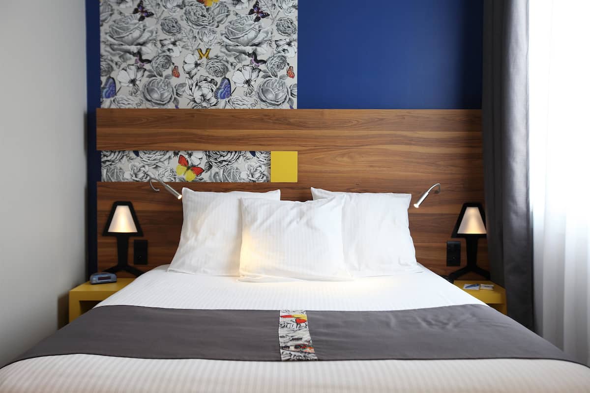 A neatly made bed with white linens, a grey bedspread, and a colorful decorative strip. There are two bedside tables with lamps, a wooden headboard, and a floral-patterned wall above the bed.