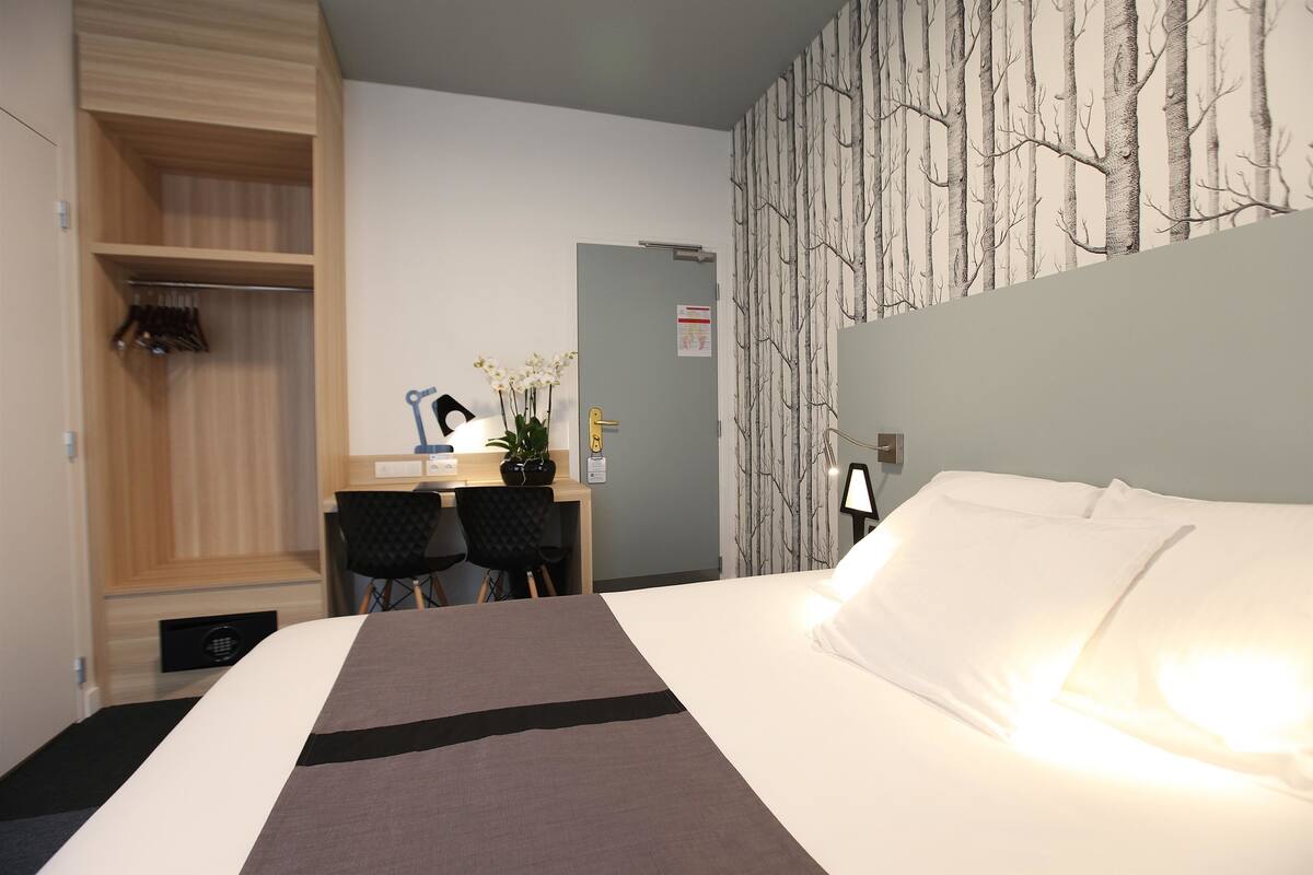 A hotel room with a bed, a desk, two chairs, a wardrobe, and a wall with a tree pattern.