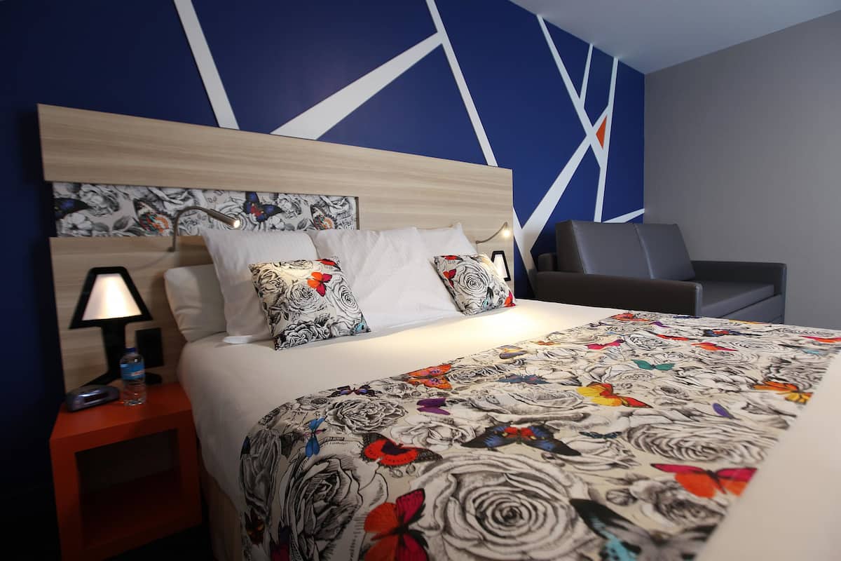 A hotel room with a bed, a nightstand with a lamp, a chair, and a wall with a geometric pattern.