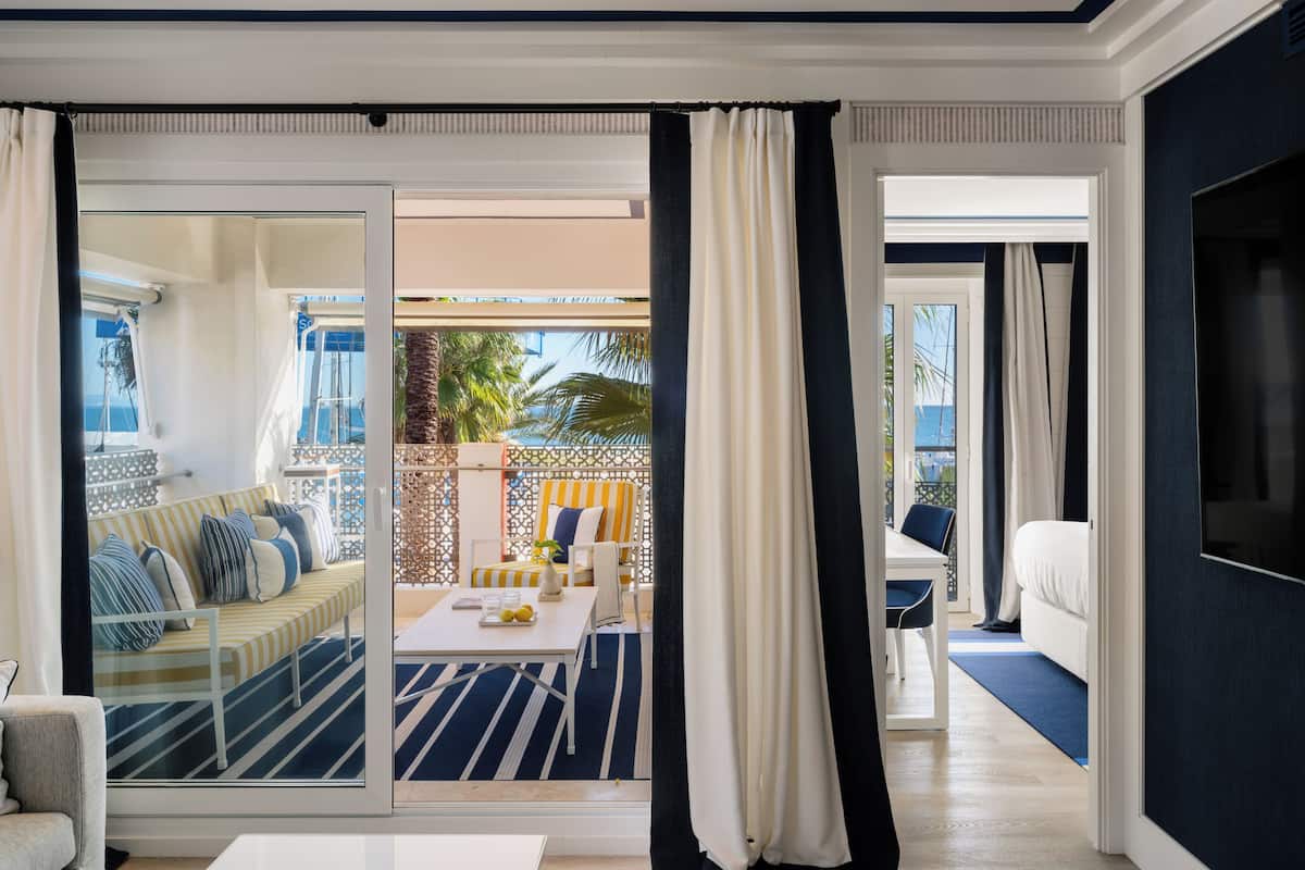 A modern hotel room with a balcony, a sofa, a dining table, and a TV.