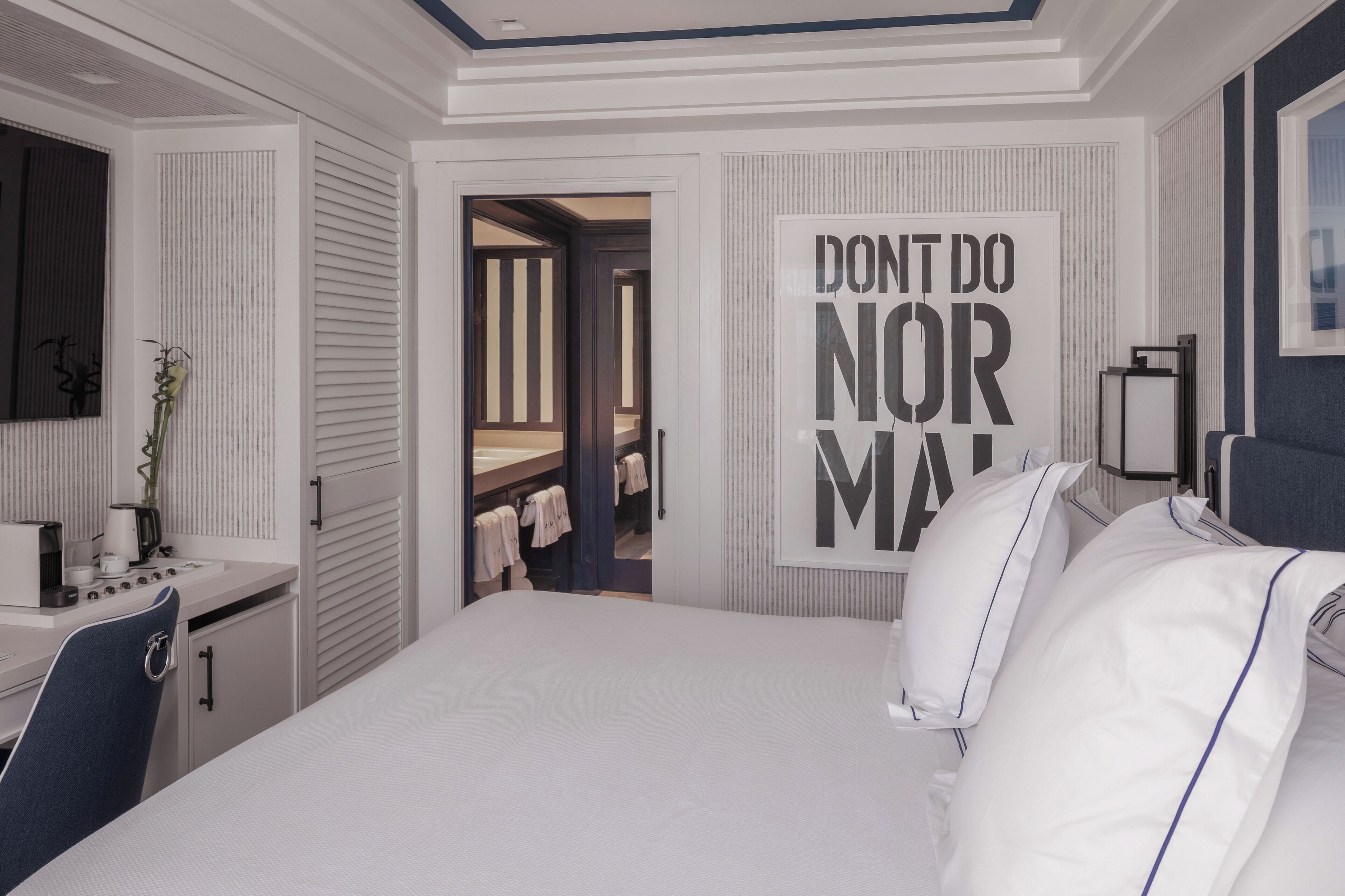 A modern hotel room with a large bed, a desk with a kettle, and a mirror. There is a door leading to a bathroom with towels hanging on the wall.