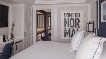 A modern hotel room with a large bed, a desk with a kettle, and a mirror. There is a door leading to a bathroom with towels hanging on the wall.
