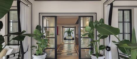 A hallway with potted plants, a patterned rug, and a view into a room with a bed and a plant.