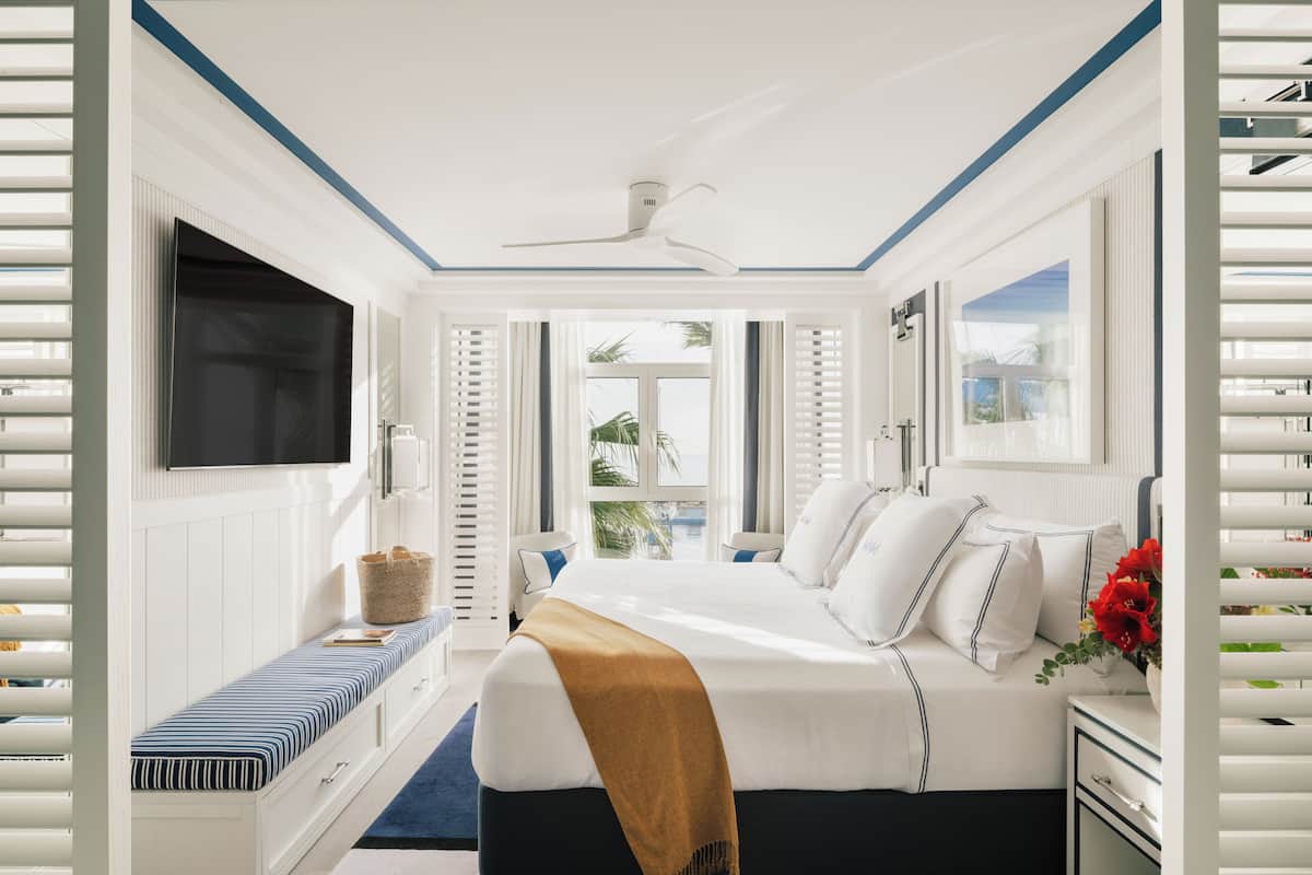 A bedroom with a large bed, a TV mounted on the wall, a window with blinds, and a ceiling fan.