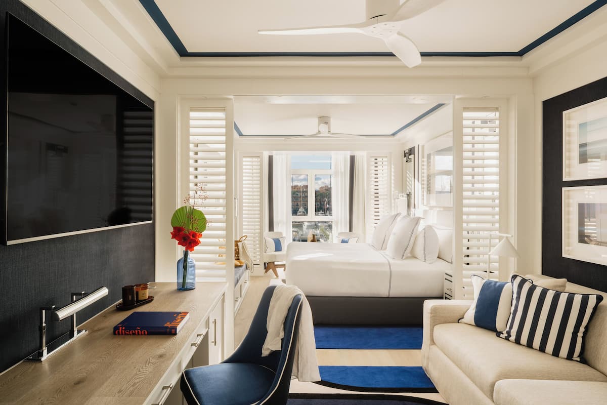 A modern hotel room with a large flat-screen TV, a desk with a book, a chair, a bed with a striped pillow, and a view of the city through the window.