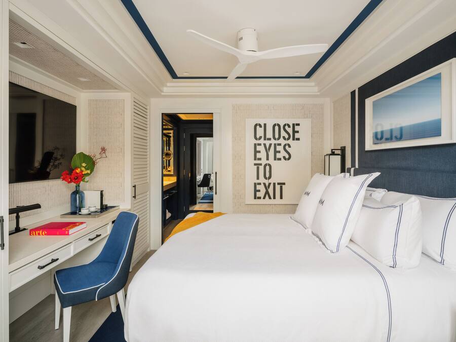 A hotel room with a large bed, a desk with a chair, a mirror, a framed artwork, and a 'CLOSE EYES TO EXIT' sign.