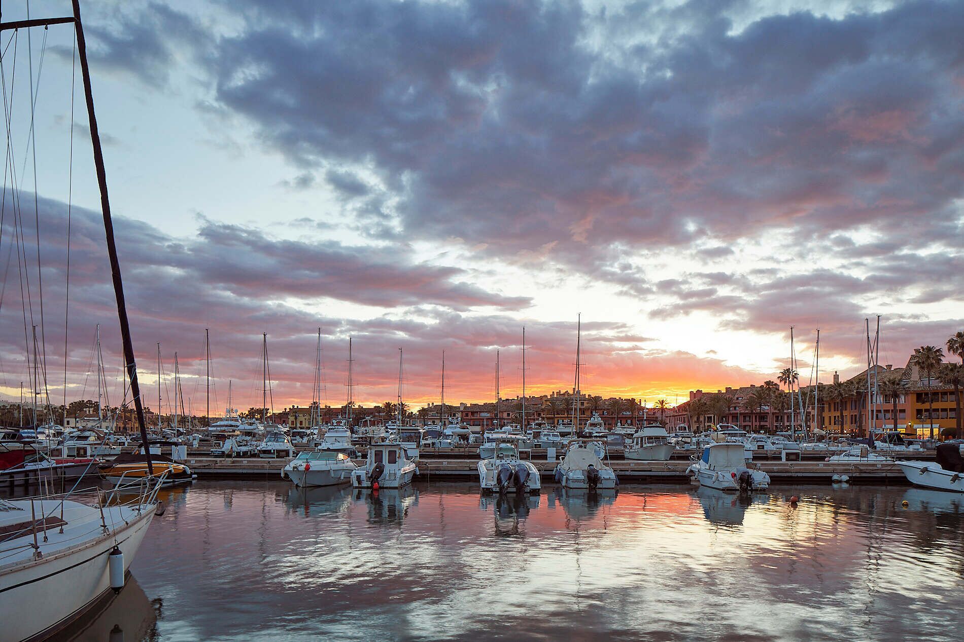 Un puerto deportivo con varias embarcaciones amarradas, un atardecer al fondo y edificios a lo largo de la costa.
