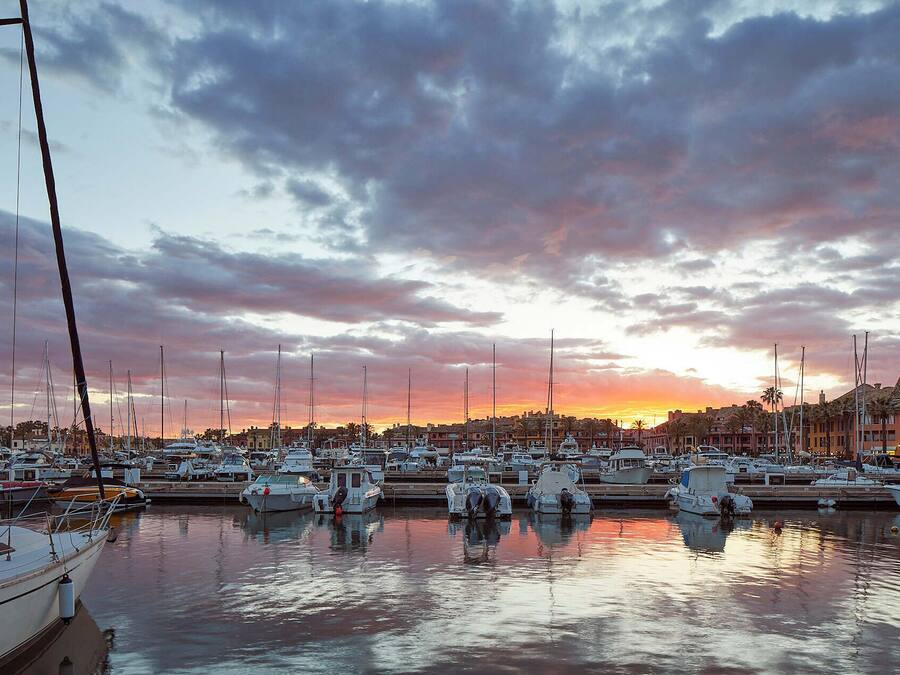 Un puerto deportivo con varias embarcaciones amarradas, un atardecer al fondo y edificios a lo largo de la costa.