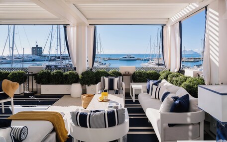 A modern, well-lit balcony with a view of a marina and boats. There are white sofas, a dining table, and potted plants.