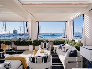 A modern, well-lit balcony with a view of a marina and boats. There are white sofas, a dining table, and potted plants.