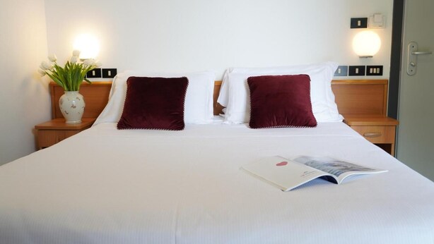 A neatly made bed with white linens and two dark red pillows, a white vase with green plants on a wooden nightstand, and a magazine on the bed. - Sunset (Rimini)