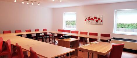 A conference room with a long table, red chairs, and a projector. There are two windows with blinds, a radiator, and a painting on the wall.