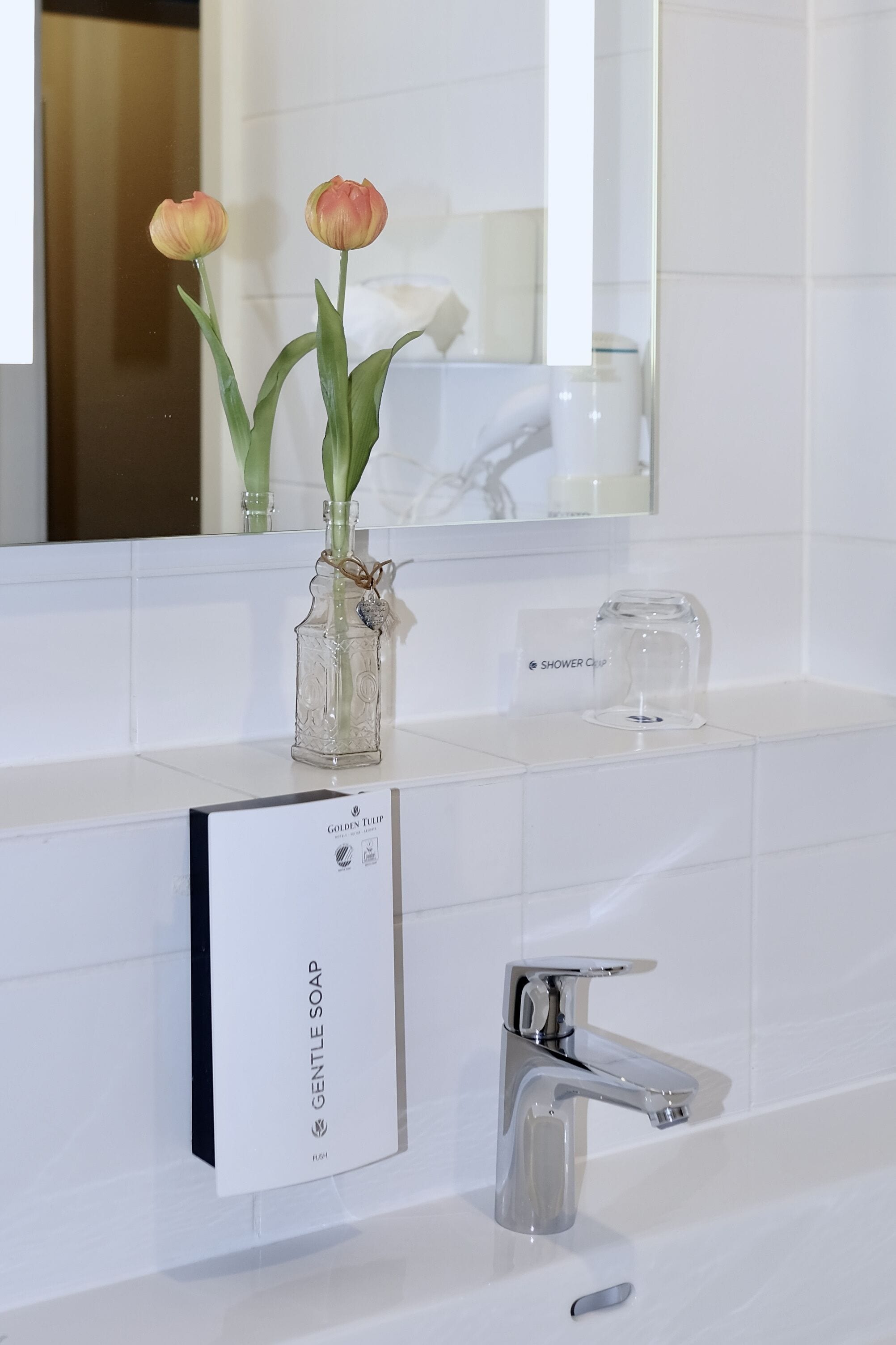 A bathroom with a white vanity, a glass vase with tulips, a bottle of shower gel, and a modern faucet.