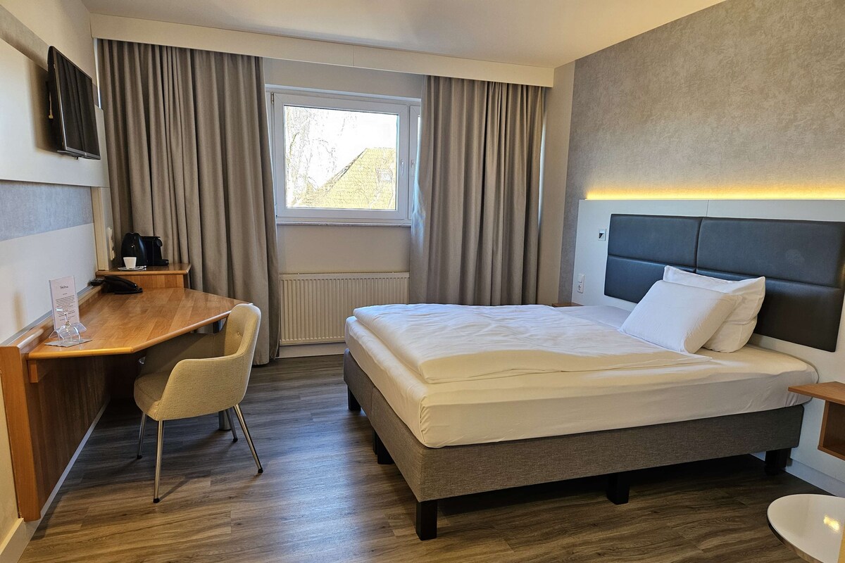 Una habitació d'hotel amb un llit, un escriptori, una cadira, una televisió i una finestra amb cortines.