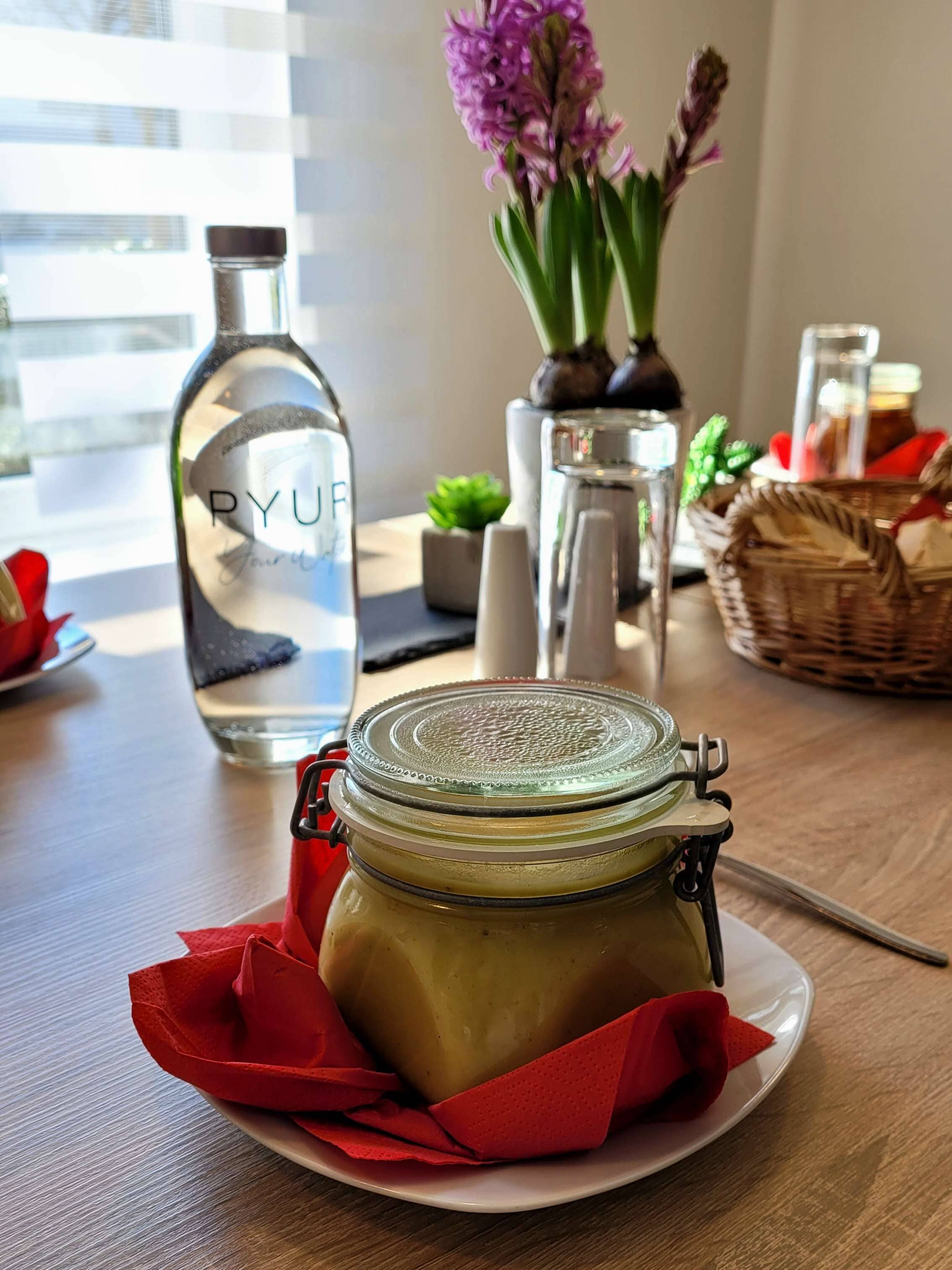 Ein Tisch mit einem Gurkarglas auf einem Teller, einer Wasserflasche, einer Blumenvase und einem Korb.