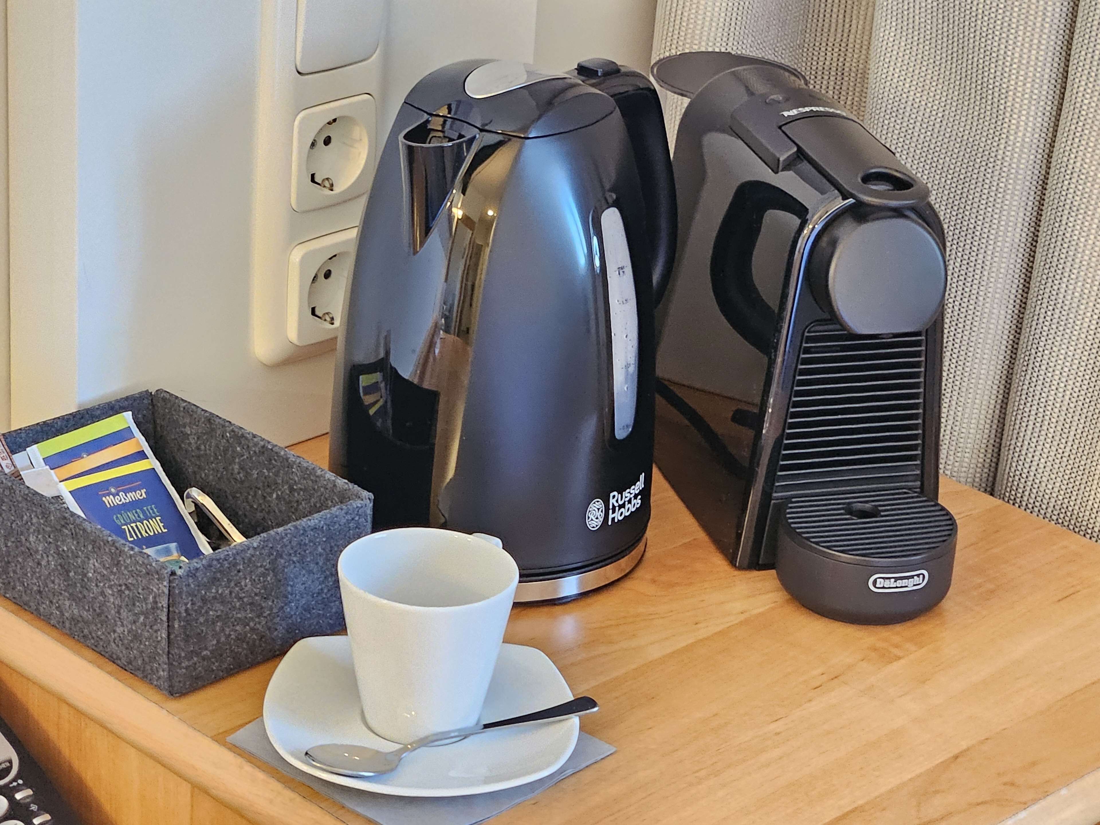 A coffee maker, a kettle, a cup, a spoon, and a box of tissues on a wooden table.
