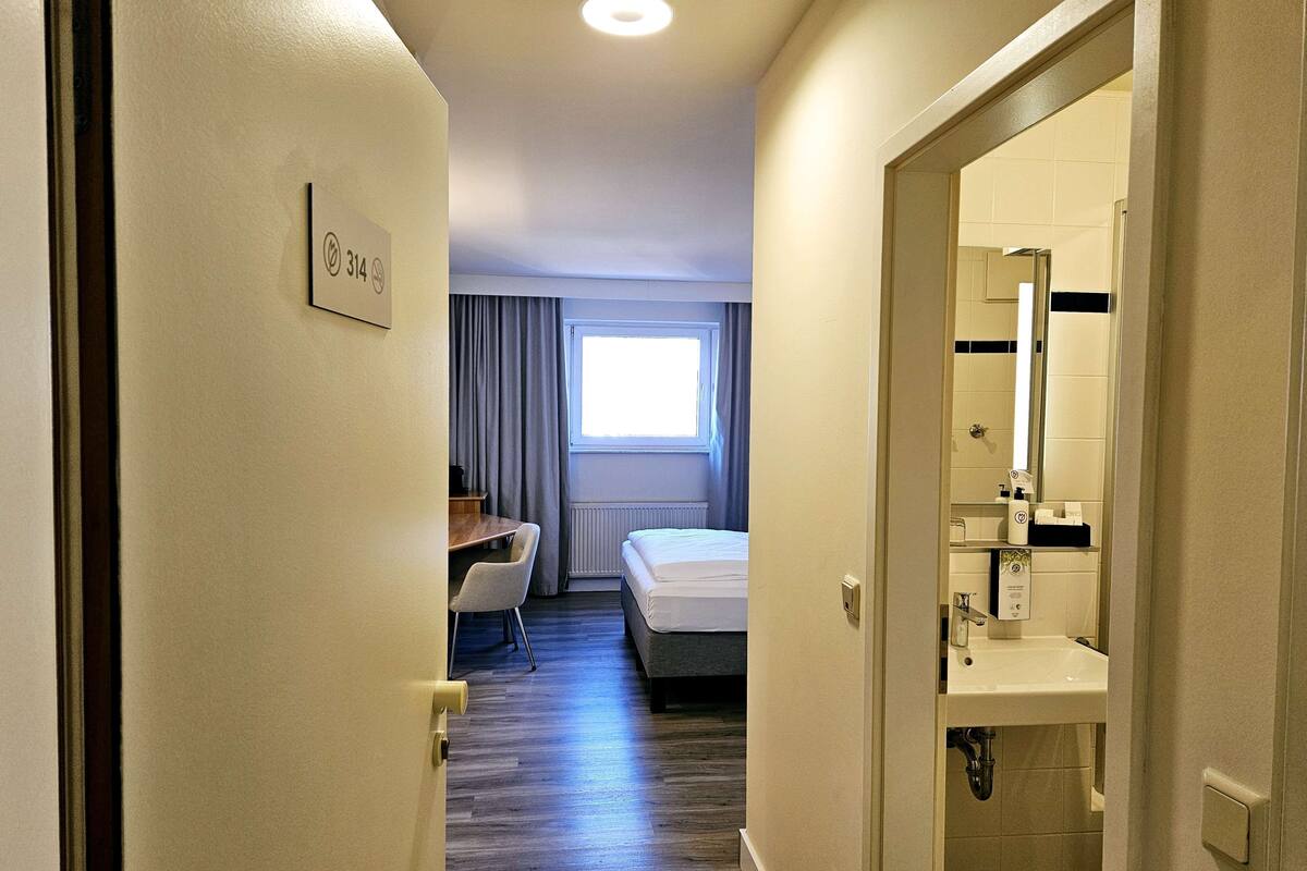 Hotelkamer met een bed, bureau, stoel, raam met gordijnen en een badkamer met wastafel en spiegel.