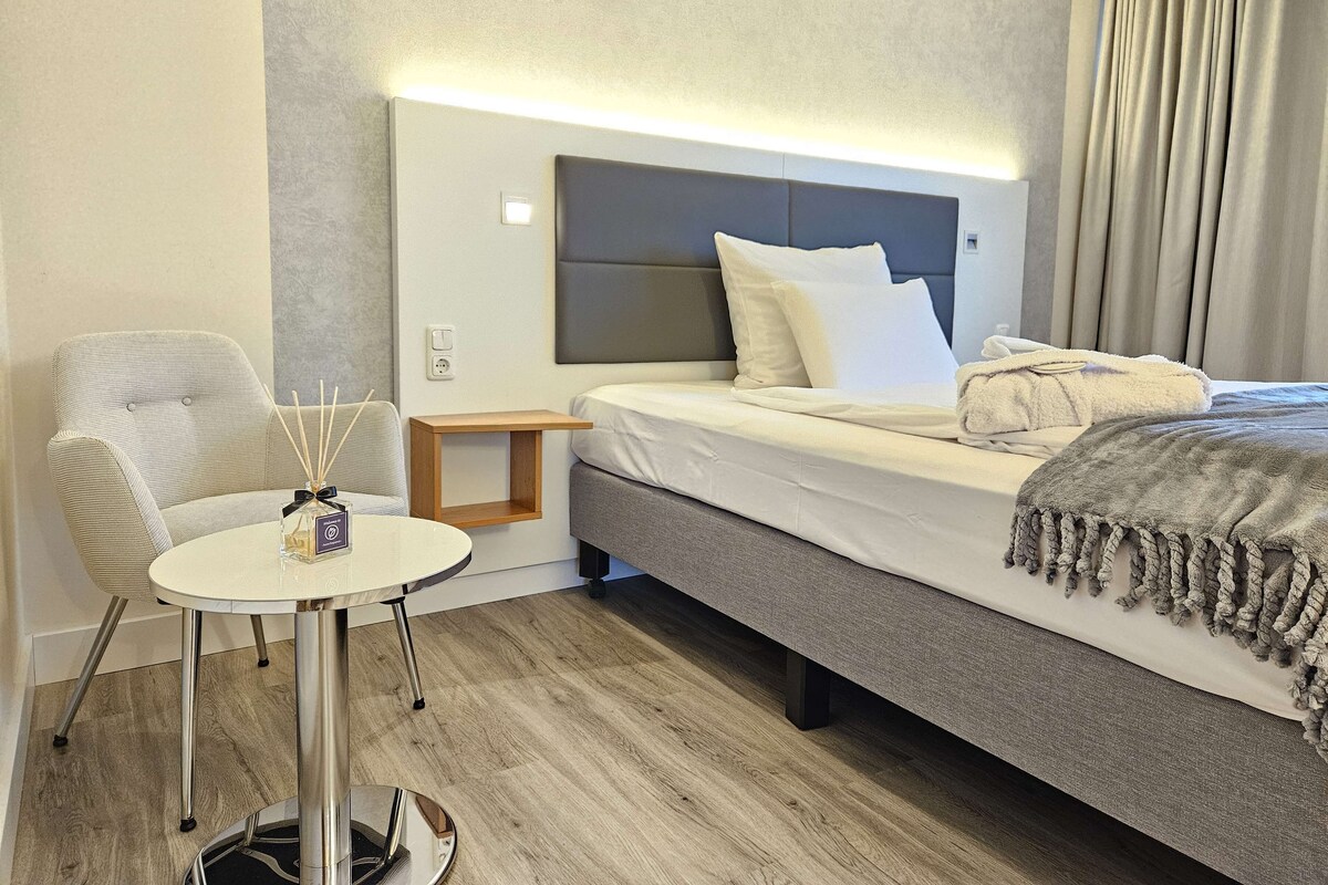 Una habitació d'hotel moderna amb un llit, una taula petita, una cadira i una taula de nit amb una ampolla d'aigua.
