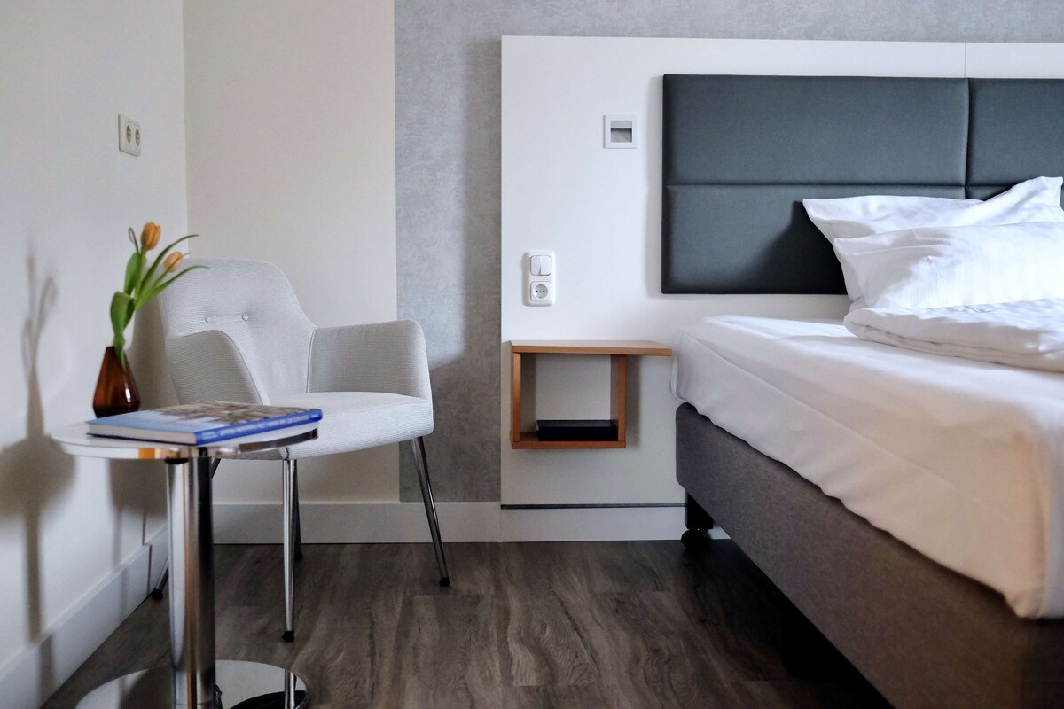 Habitació d'hotel moderna amb un llit, una petita taula de nit, una cadira i una prestatgeria penjada a la paret.