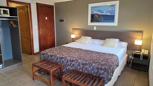 Ein Hotelzimmer mit einem Bett, zwei Nachttischlampen, einer Holzbank, einem Kleiderschrank und einem Gemälde einer Berglandschaft.