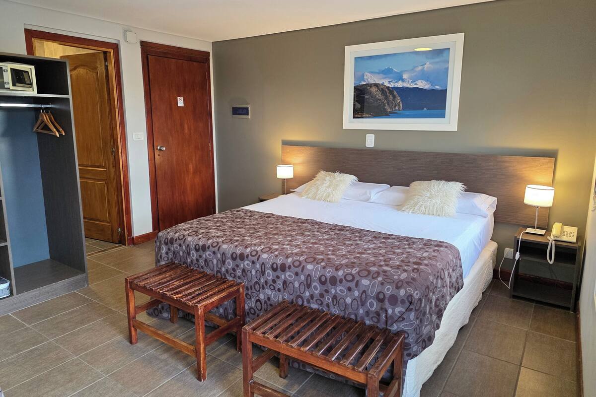 Une chambre d’hôtel comprenant un lit, deux lampes de chevet, un banc en bois, une armoire et un tableau représentant un paysage de montagne.