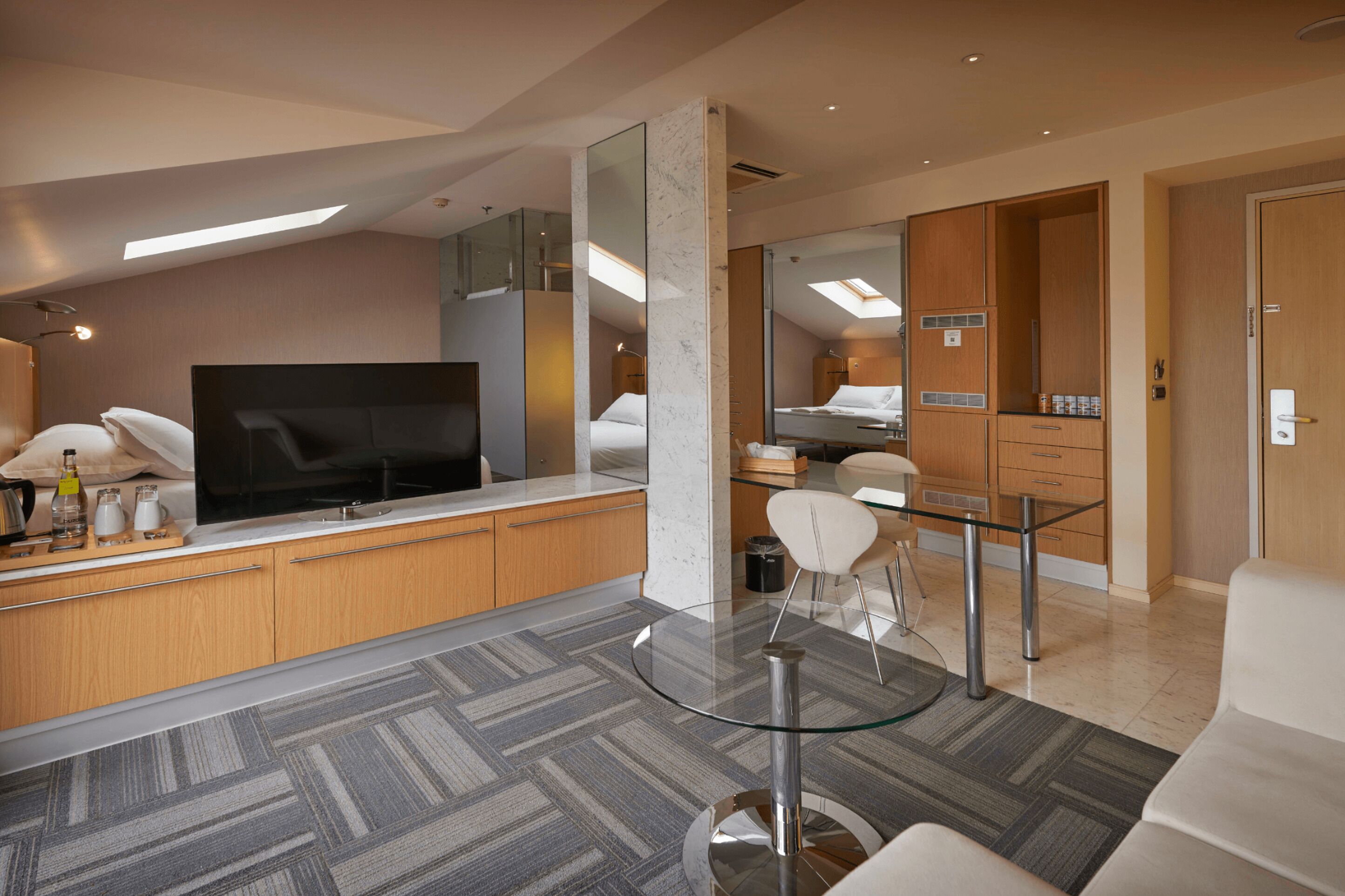 A modern hotel room with a large flat-screen TV, a glass-top dining table, a sofa, and a large mirror.