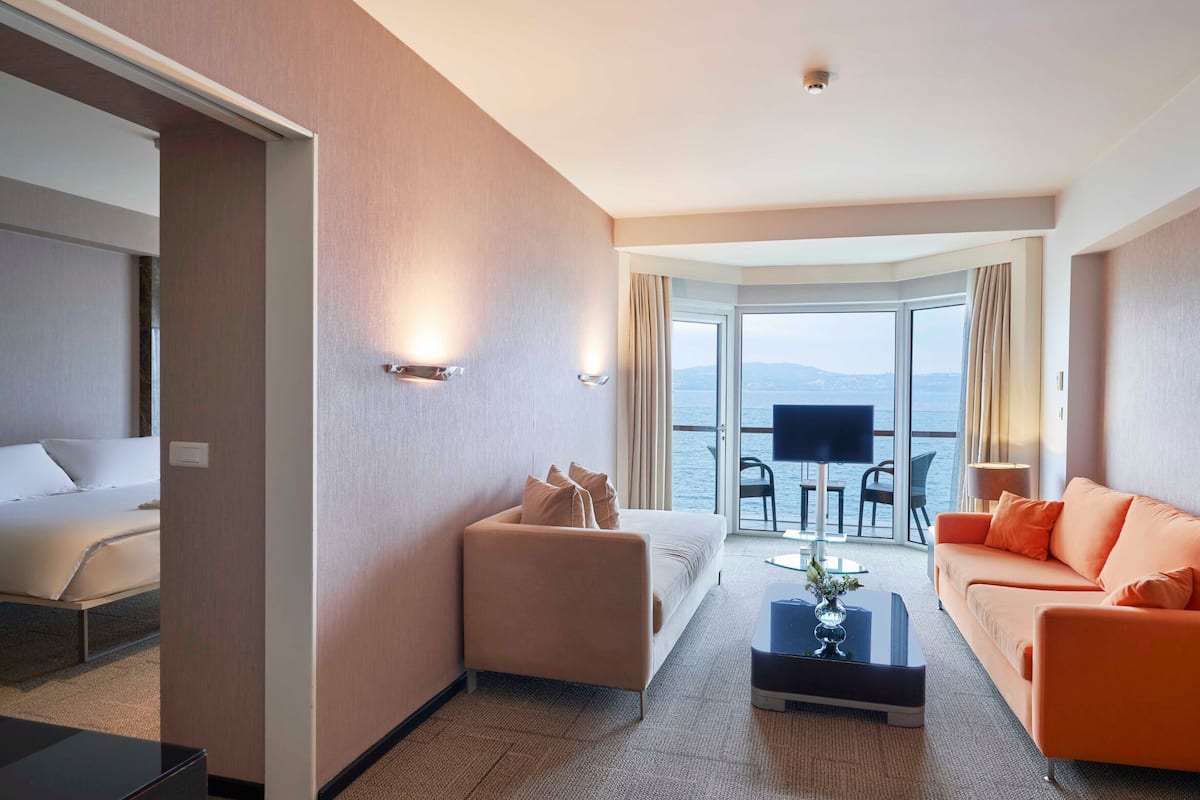 A hotel room with a bed, a sofa, a coffee table, and a TV. There is a large window with a view of the sea.