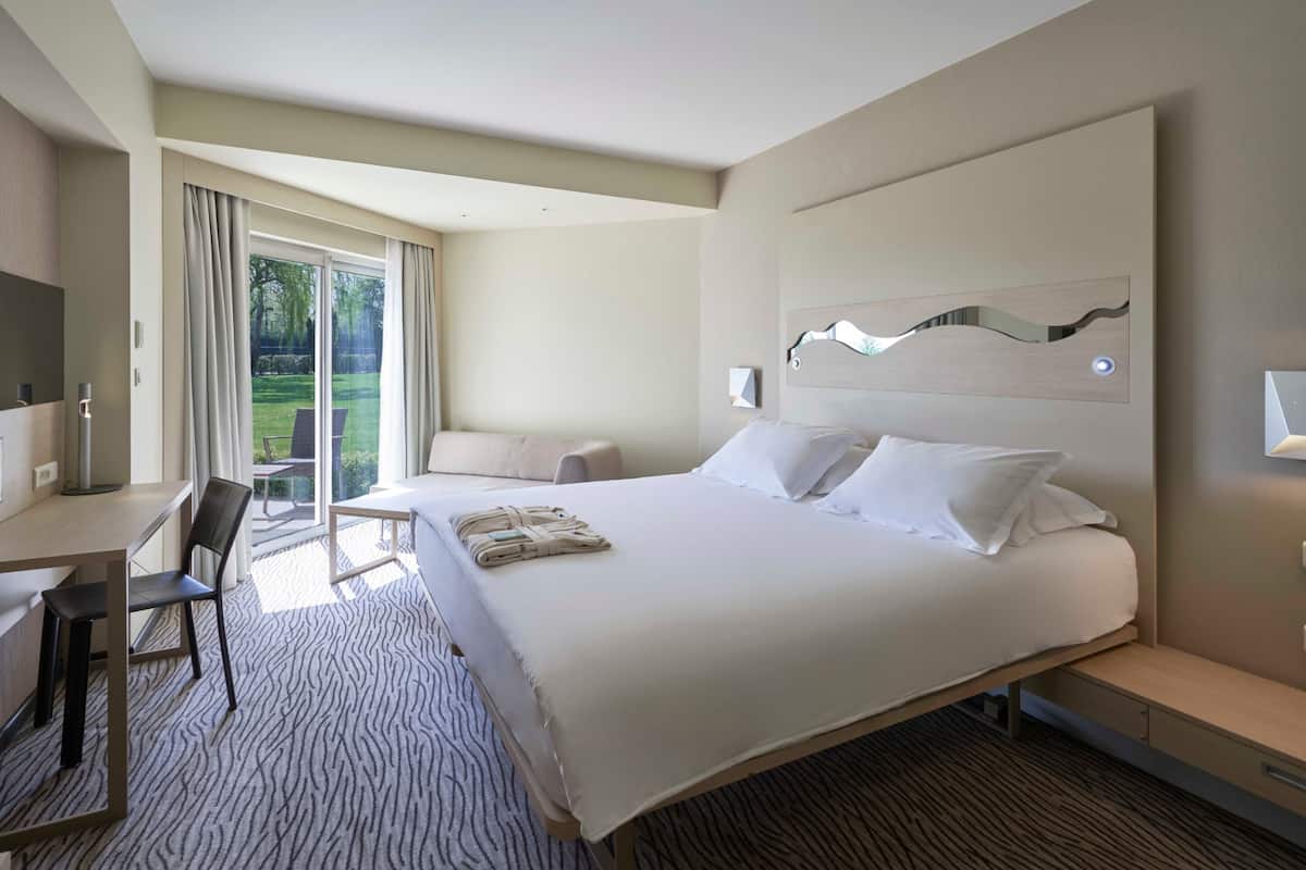 A modern hotel room with a large bed, a desk, and a chair. There is a view of a garden through the window.