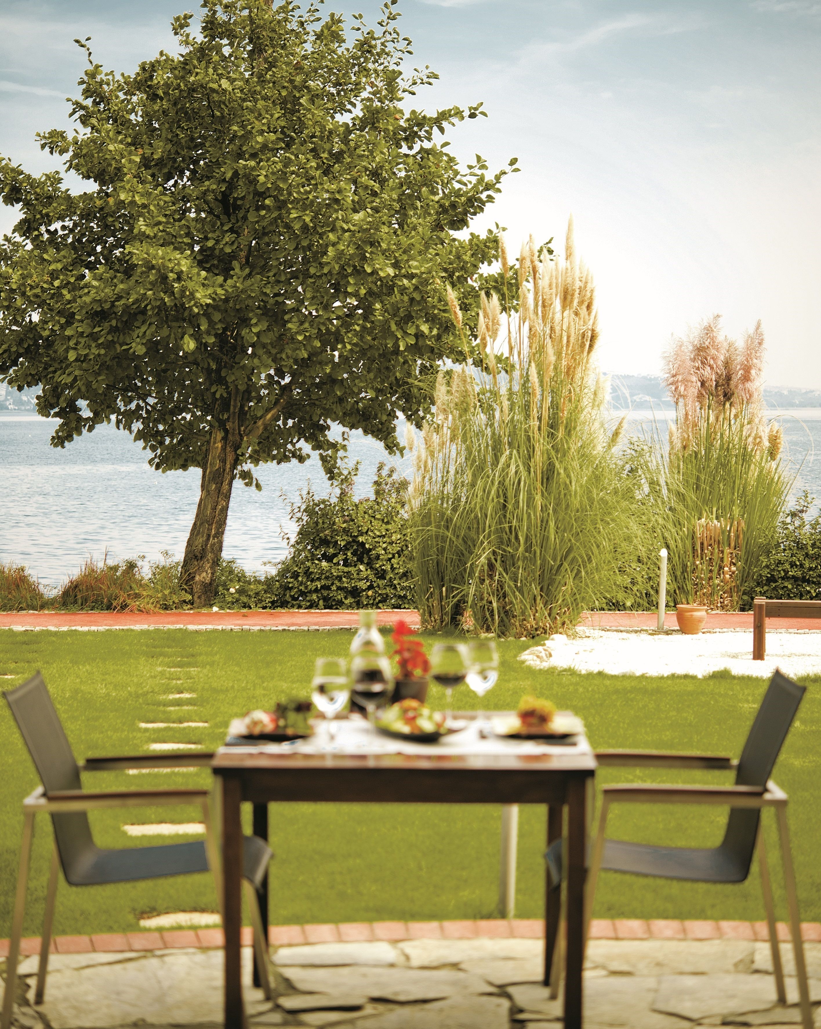 Un área de comedor al aire libre con una mesa para dos, rodeada de sillas, con vista a un cuerpo de agua y un árbol grande.