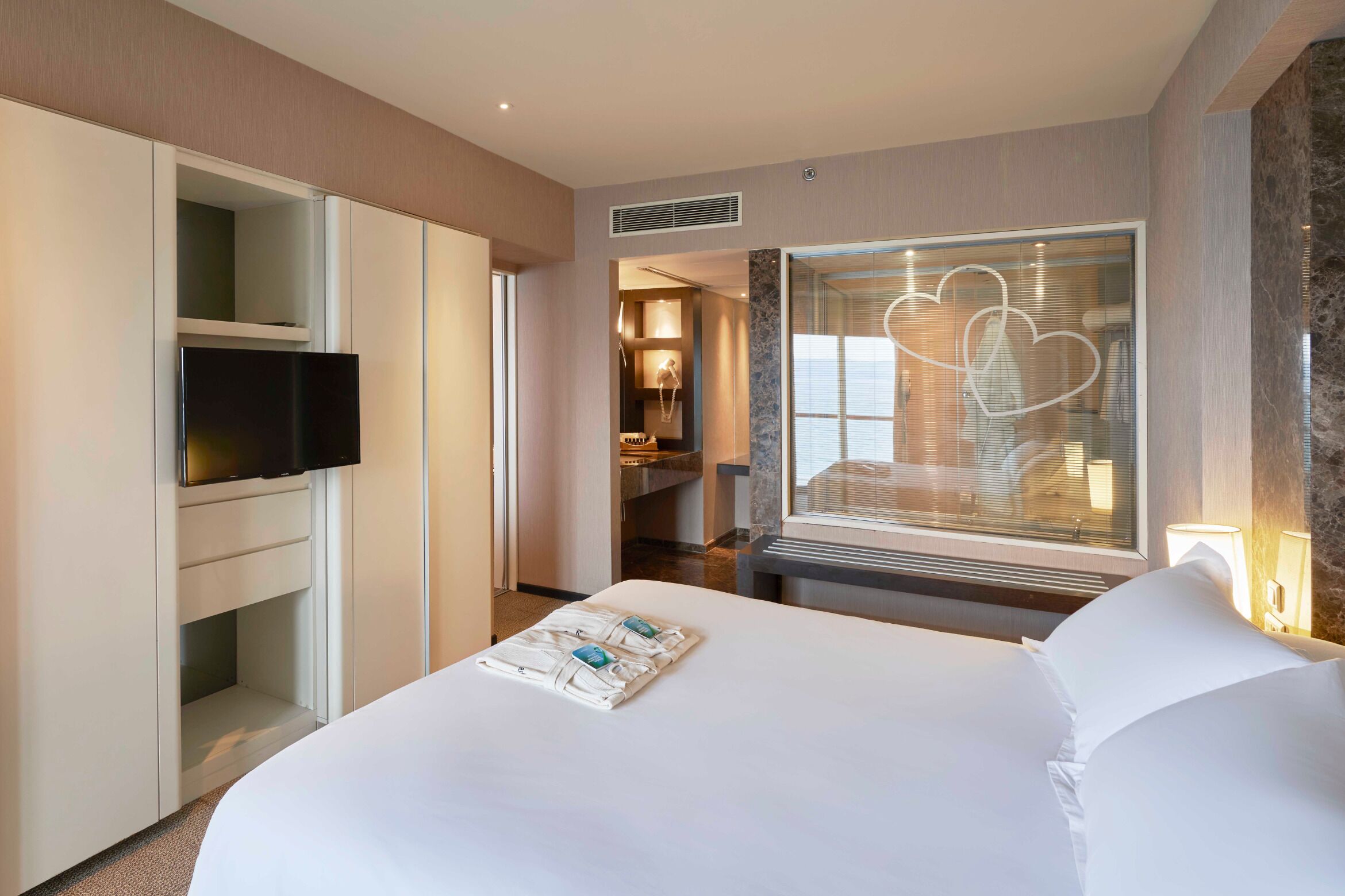 Ein Hotelzimmer mit einem großen Bett, einem an einer Kommode befestigten Fernseher, einem Badezimmer mit Spiegel und einem Fenster mit Herzmotiv.