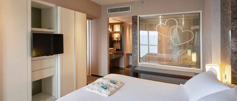 Ein Hotelzimmer mit einem großen Bett, einem an einer Kommode befestigten Fernseher, einem Badezimmer mit Spiegel und einem Fenster mit Herzmotiv.