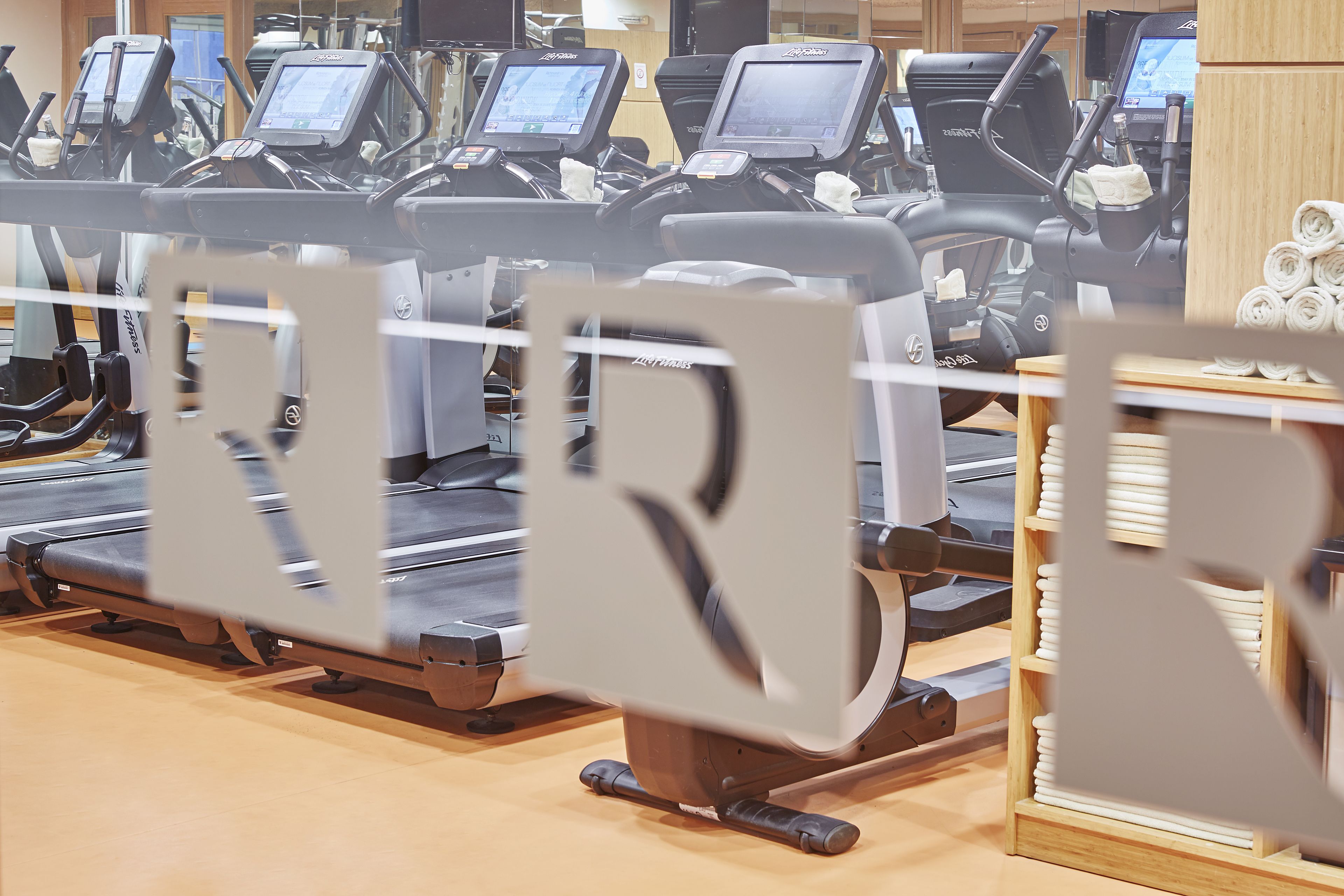 A row of stationary bikes with digital screens, a wooden floor, and a towel rack in the background.